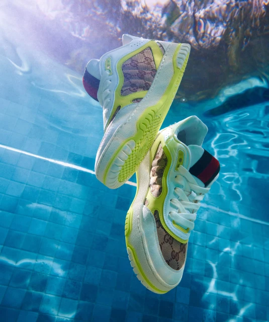 Elevated view of two white Gucci sneakers floating in a blue-tiled swimming pool with underwater lighting creating a surreal fashion image.