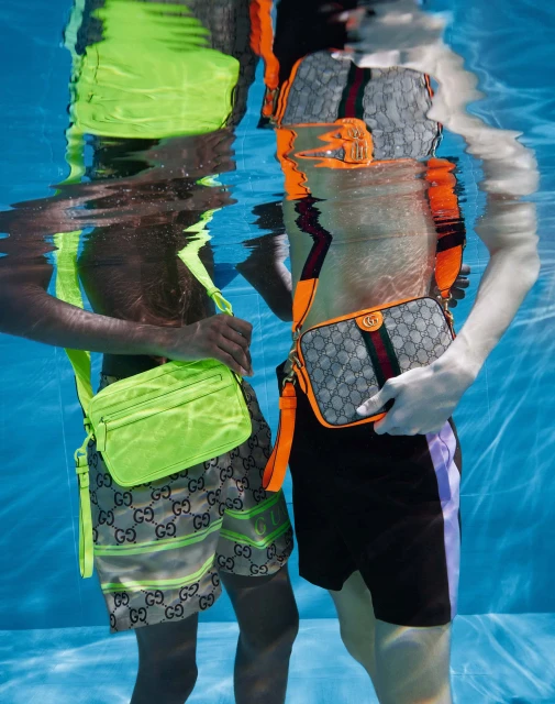 Two people in a pool underwater, showcasing Gucci bags and matching Gucci patterned swimwear. The bags are bright neon and muted grey.