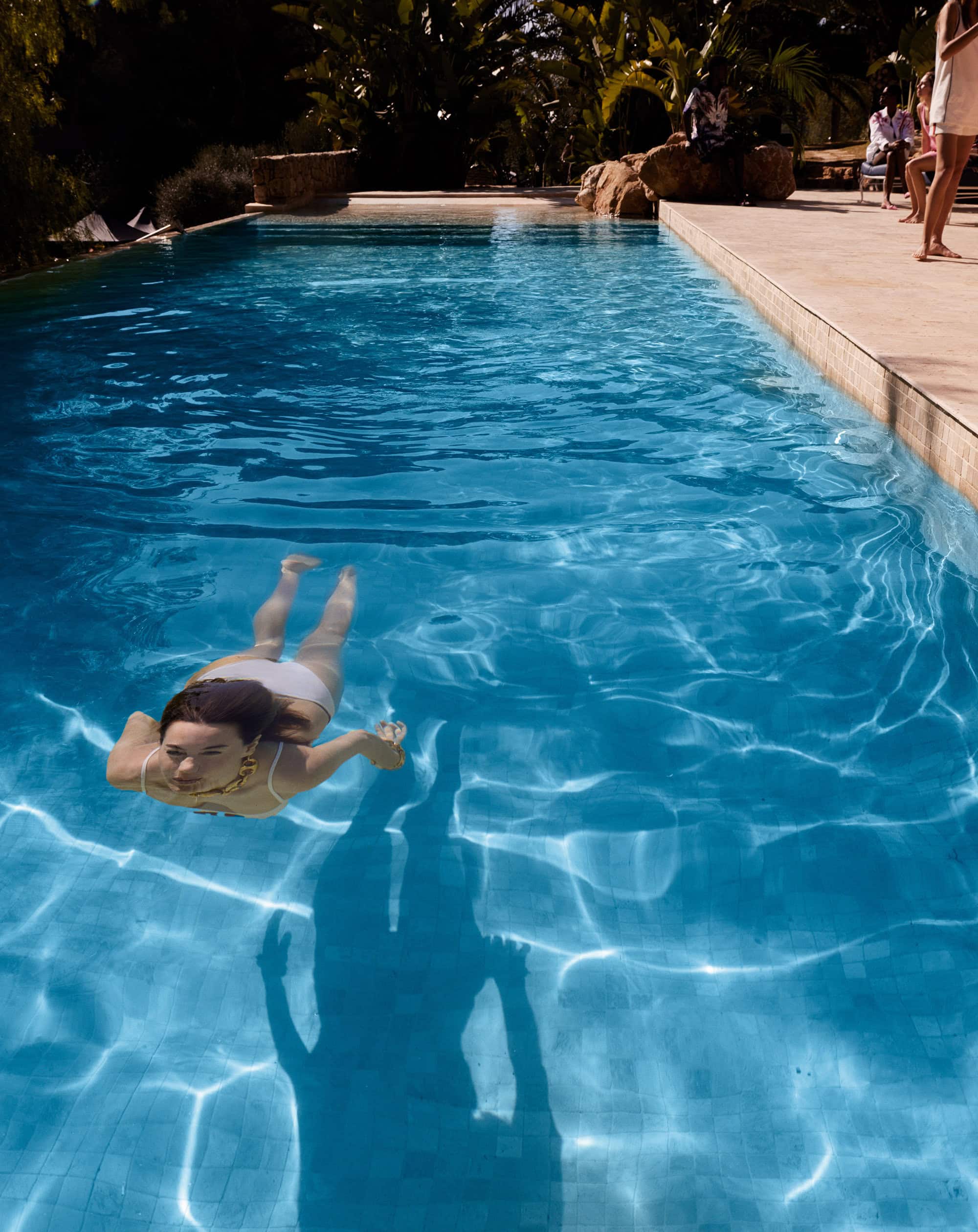 Woman in a white swimsuit swims in a bright turquoise outdoor swimming pool with sunlight reflecting off the water.