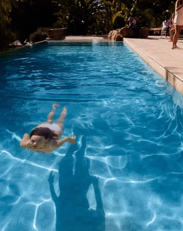 Woman in a white swimsuit swims in a bright turquoise outdoor swimming pool with sunlight reflecting off the water.