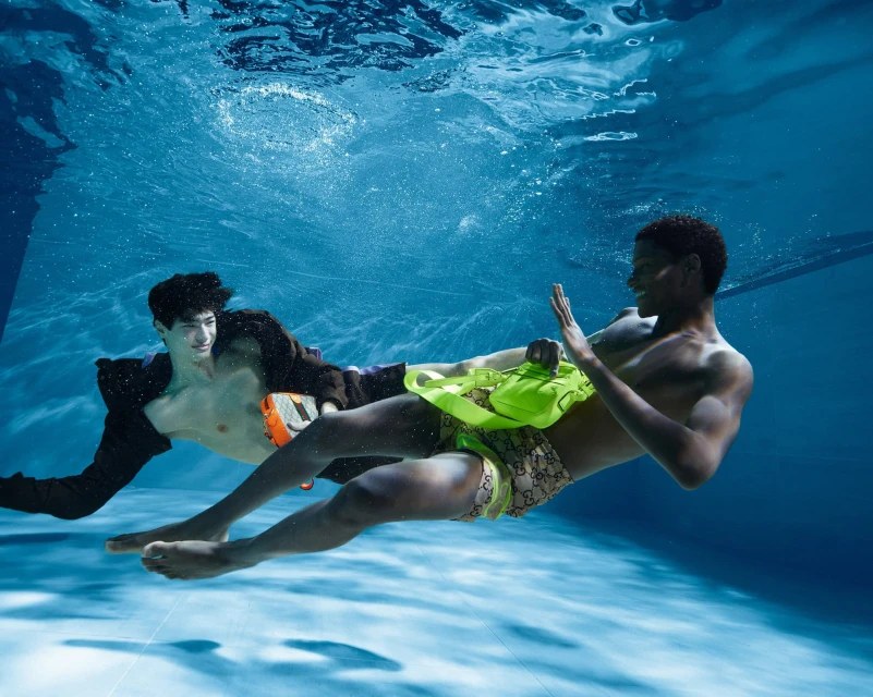 Two young men in swimming trunks are underwater, one in a black jacket, in a pool of clear blue water with tiled floor.
