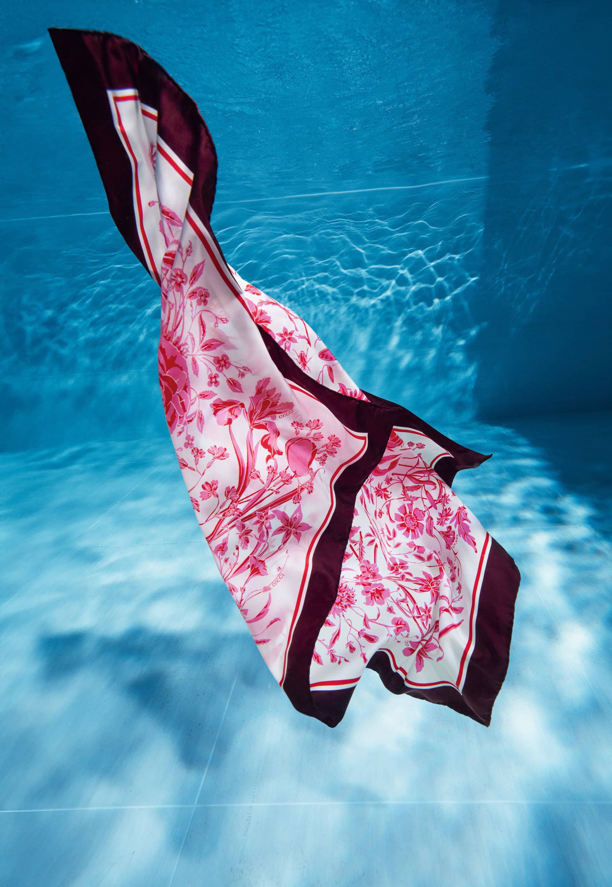 Floral patterned silk scarf with a burgundy border floats underwater in a blue-toned pool with sunlight creating dappled patterns.