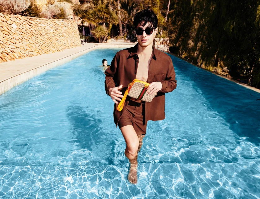 Man wading in a pool holding a handbag, wearing sunglasses and a brown shirt and shorts, with stone wall and palms in background.