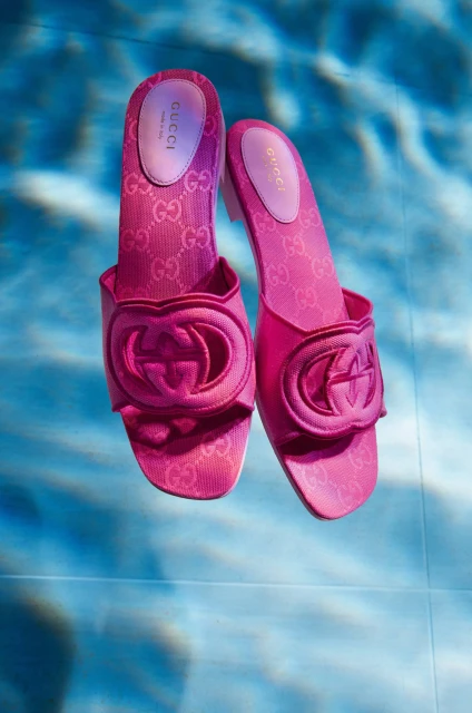 Pair of pink Gucci slide sandals with the brand's logo, showcased against a textured blue backdrop suggesting a pool or water surface.