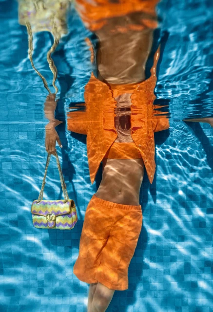 Underwater fashion shot featuring a woman in an orange suit holding two handbags in a blue-tiled swimming pool with light rays.