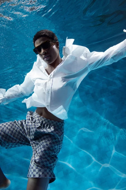 An adult swims underwater, wearing sunglasses, a white button-down shirt and patterned shorts, against a blue water background.