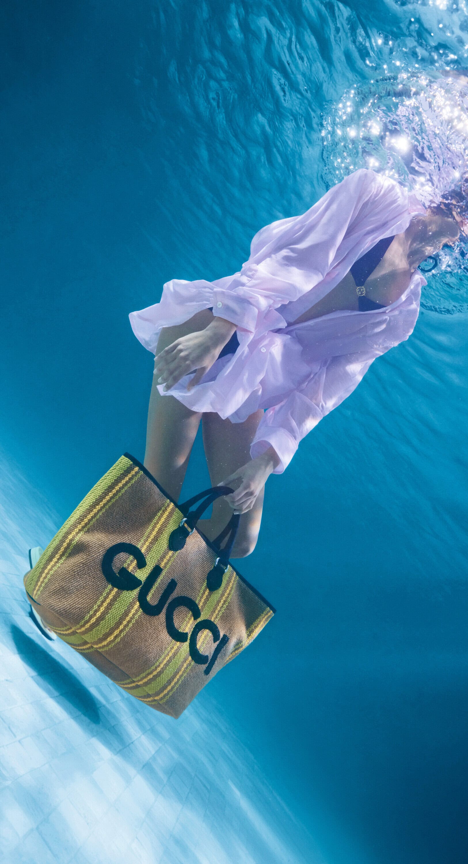 Underwater fashion shot of a woman in a white shirt holding a Gucci bag in a blue underwater setting with visible sunlight.