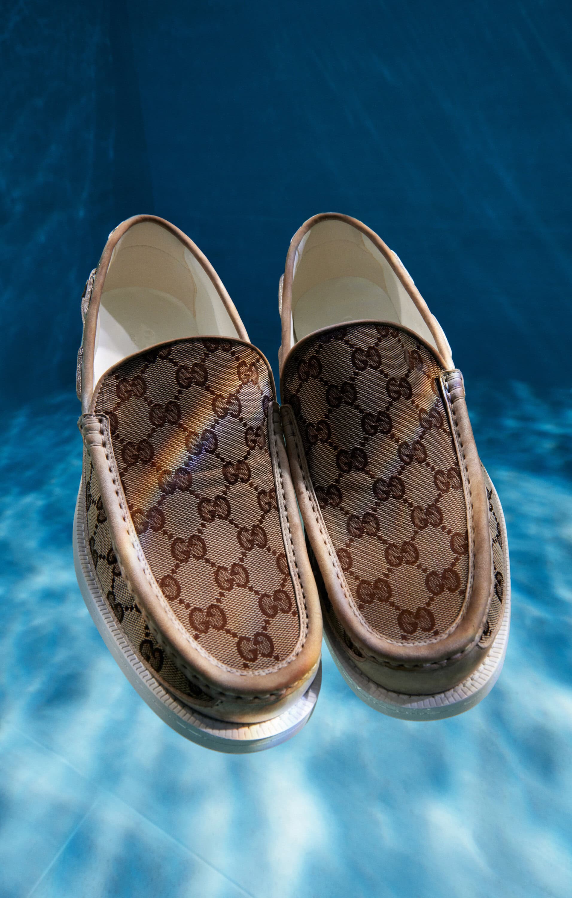 Two Gucci slip-on shoes with a brown logo pattern are displayed against a blue, watery background, creating a visually striking effect.