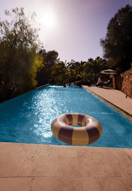 Swimming pool view featuring a striped inflatable ring in the water, with a villa, trees and other people in the background on a sunny day.