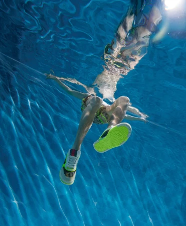 Underwater shot of a person in a swimming pool wearing sneakers, captured at eye-level with natural light filtering through the water.