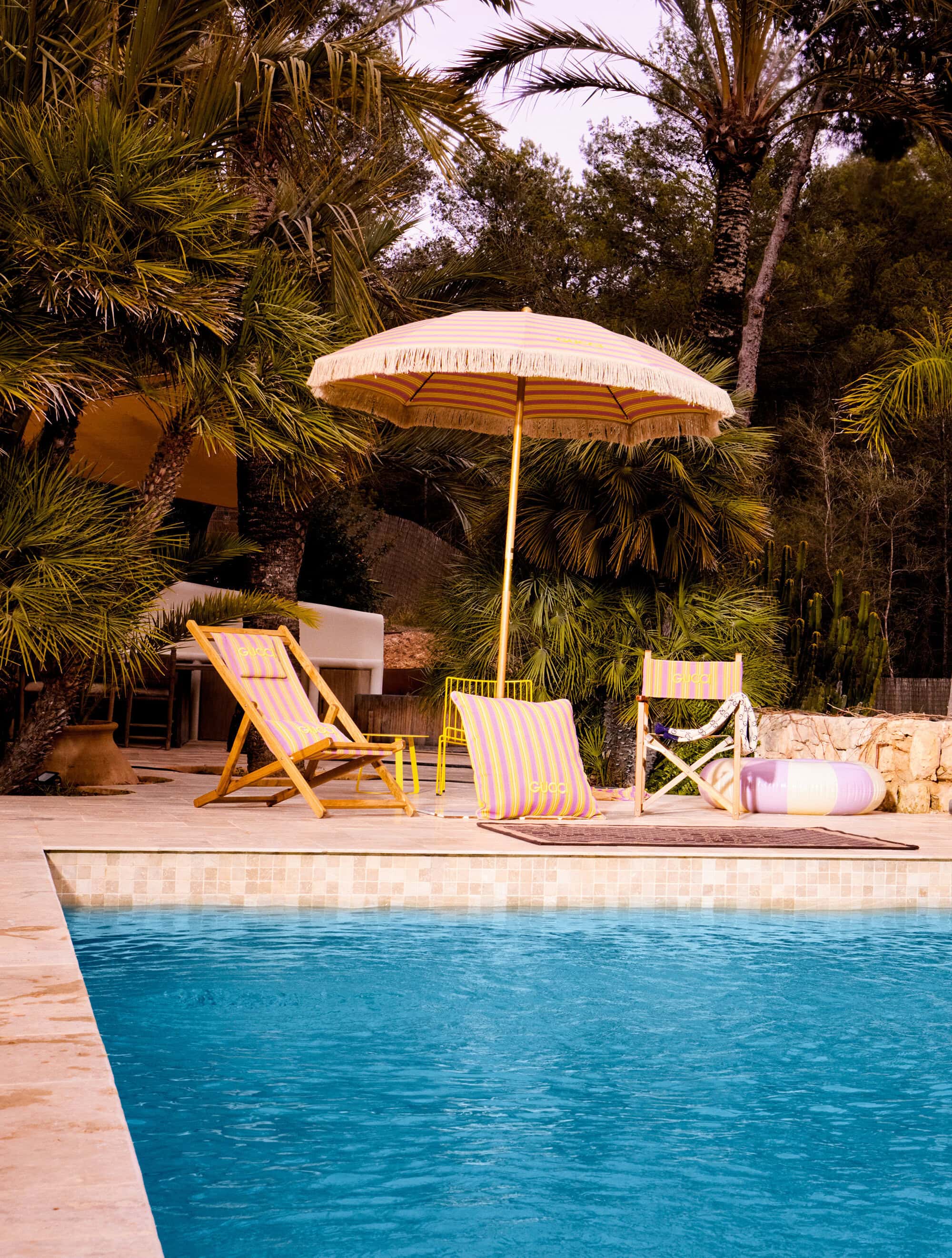 Outdoor poolside setting with a sun umbrella, lounge chair, and beach accessories next to a turquoise swimming pool surrounded by lush foliage.