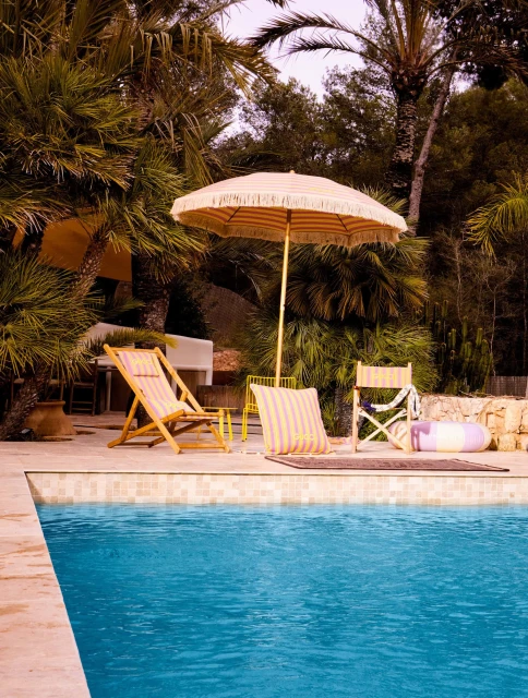 Outdoor poolside setting with a sun umbrella, lounge chair, and beach accessories next to a turquoise swimming pool surrounded by lush foliage.