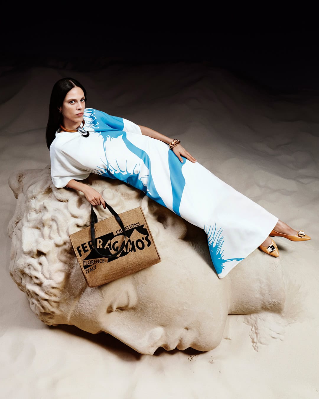 Model in blue and white printed dress reclining on a giant stone head sculpture holding a beige handbag