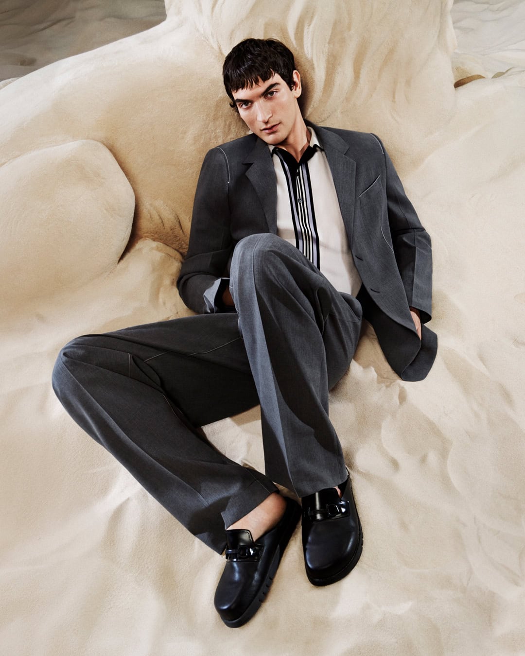 A man in a grey suit and black loafers is reclining on a light-beige surface. He is wearing a striped shirt. Medium shot, indoor setting.