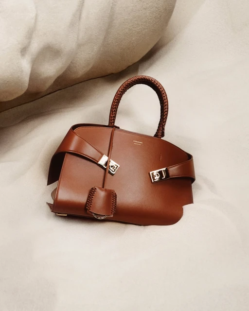 Studio shot of a brown leather handbag with a braided handle, featuring silver hardware and resting on a light-colored wrinkled fabric.