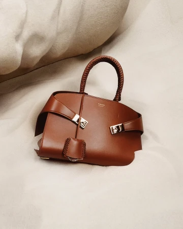 Studio shot of a brown leather handbag with a braided handle, featuring silver hardware and resting on a light-colored wrinkled fabric.