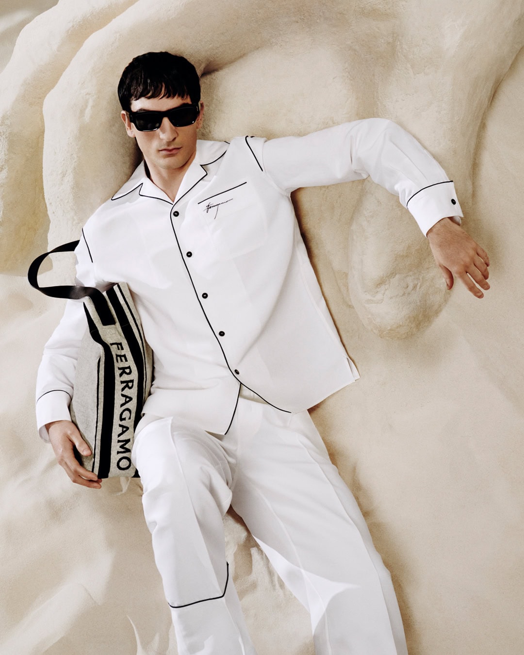 Man lies on sand wearing white pajamas and sunglasses, holding a Ferragamo tote bag in a fashion editorial layout.