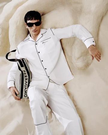 Man lies on sand wearing white pajamas and sunglasses, holding a Ferragamo tote bag in a fashion editorial layout.