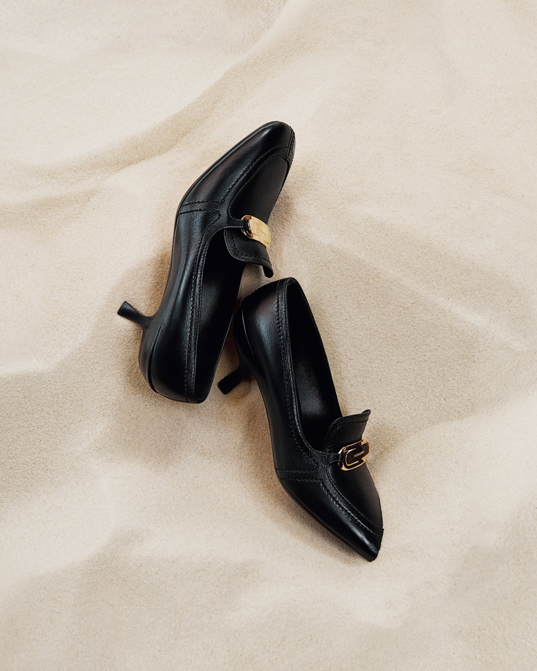Two black leather kitten heel loafers with gold hardware placed on pale sand for the Ferragamo Pre-Fall 2024 campaign