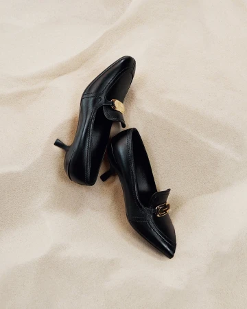 Two black leather kitten heel loafers with gold hardware placed on pale sand for the Ferragamo Pre-Fall 2024 campaign