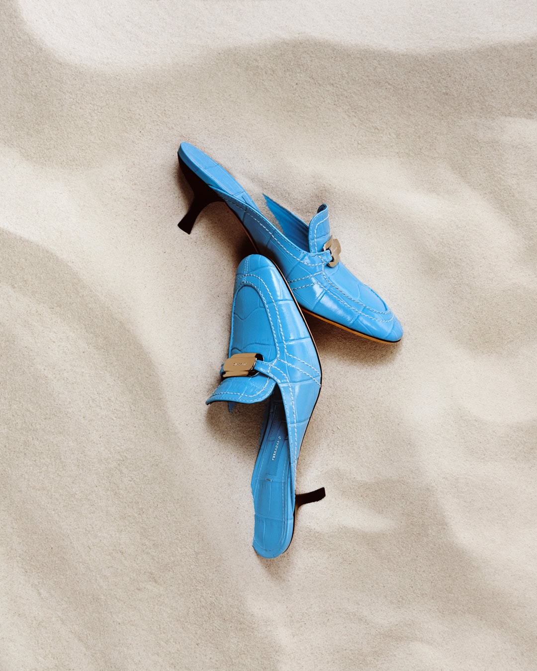 Two light-blue mules with gold hardware are displayed on a beige, textured surface in a well-lit, minimalist, and clean studio shot.