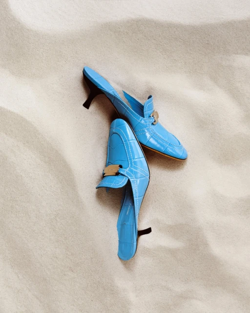 Two light-blue mules with gold hardware are displayed on a beige, textured surface in a well-lit, minimalist, and clean studio shot.
