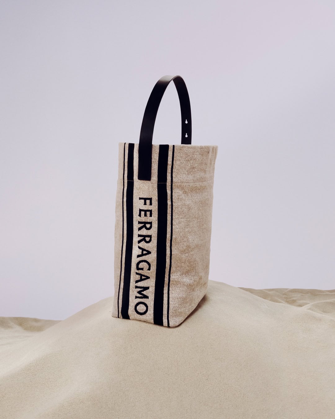 Tote bag by Ferragamo featuring beige canvas, black stripes and black leather handle, on a sand dune against a plain sky.