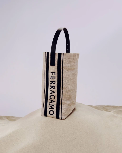Tote bag by Ferragamo featuring beige canvas, black stripes and black leather handle, on a sand dune against a plain sky.