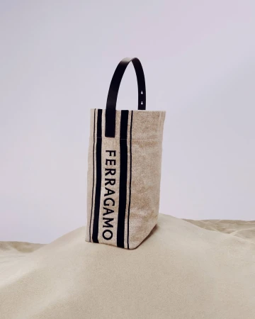 Tote bag by Ferragamo featuring beige canvas, black stripes and black leather handle, on a sand dune against a plain sky.