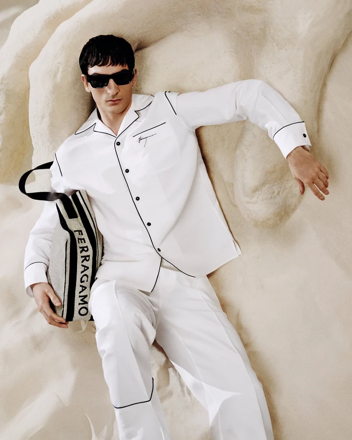 Man wearing white button-down pajama-style shirt and matching pants with black sunglasses holding a canvas tote bag