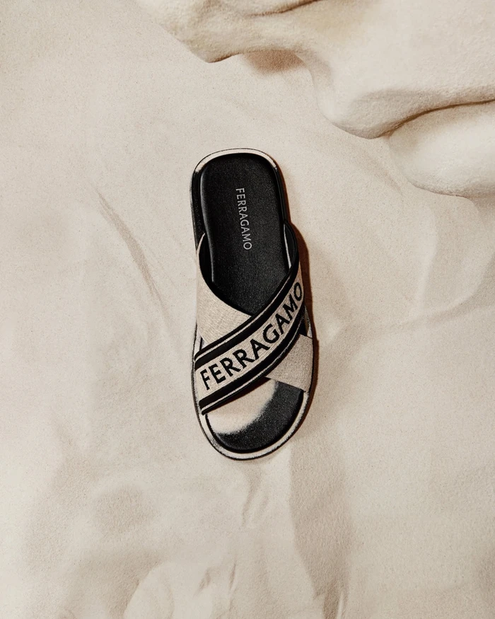 Ferragamo branded slide sandal rests on a soft sandy dune surface from an overhead perspective.