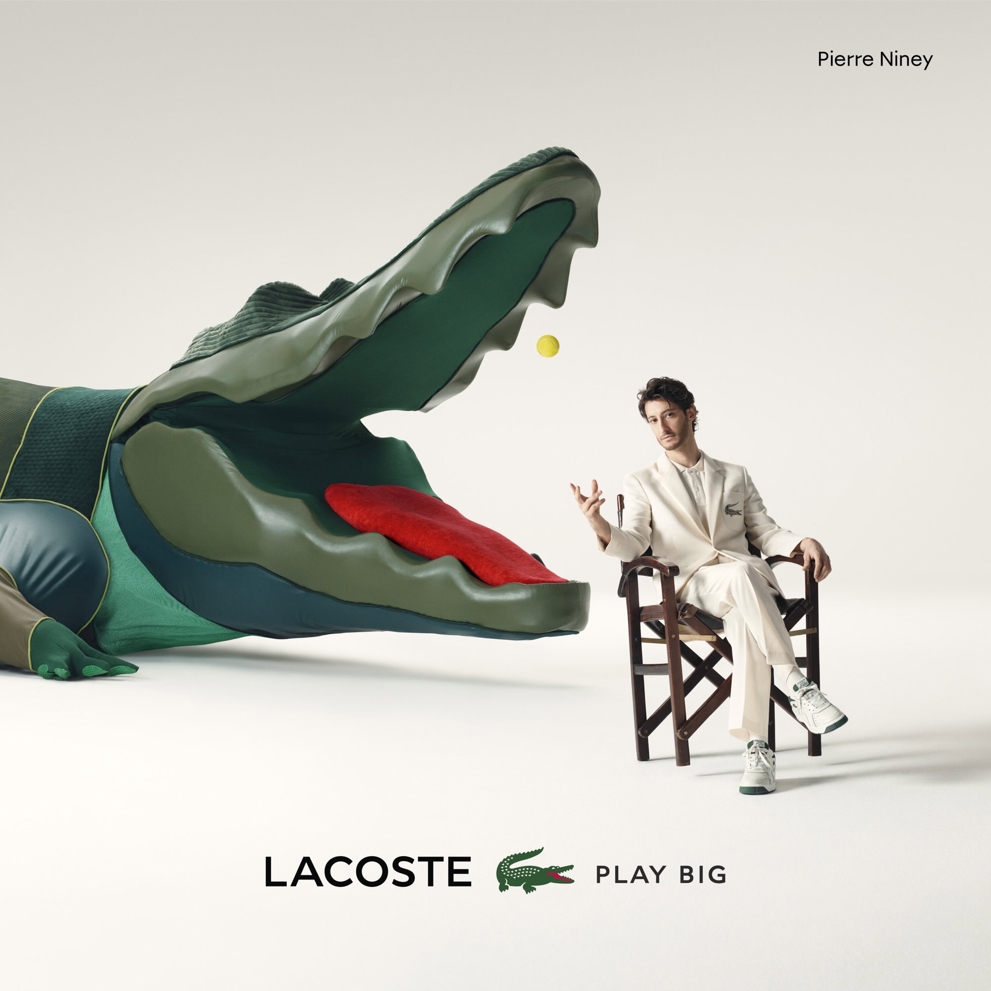 Lacoste ad featuring Pierre Niney seated by a giant crocodile head, with Lacoste logo, on a white background. Editorial style.