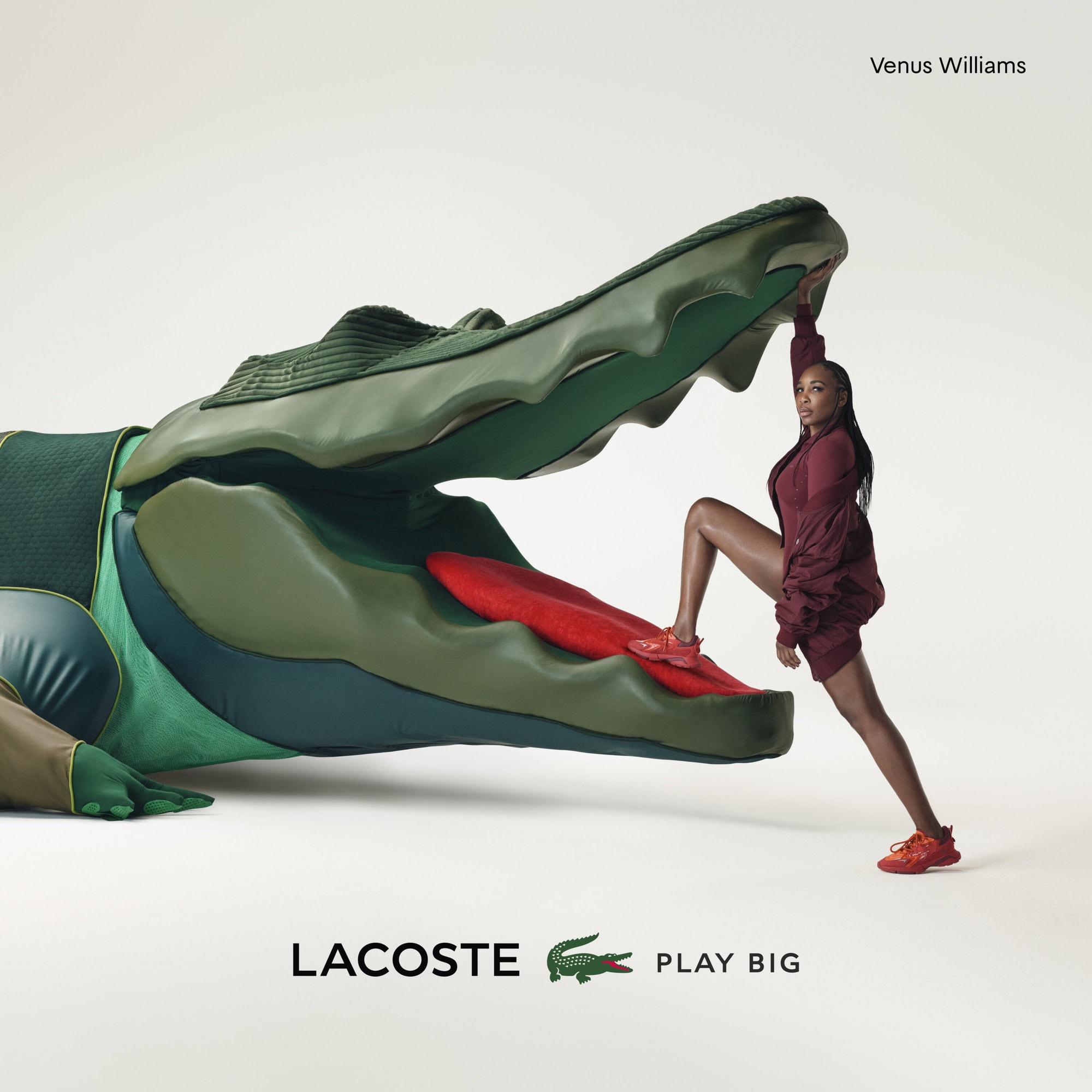 Venus Williams poses with a large Lacoste alligator prop in a studio shot, wearing a matching red outfit and sneakers for an advertisement.