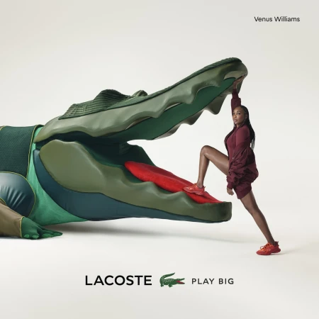 Venus Williams poses with a large Lacoste alligator prop in a studio shot, wearing a matching red outfit and sneakers for an advertisement.