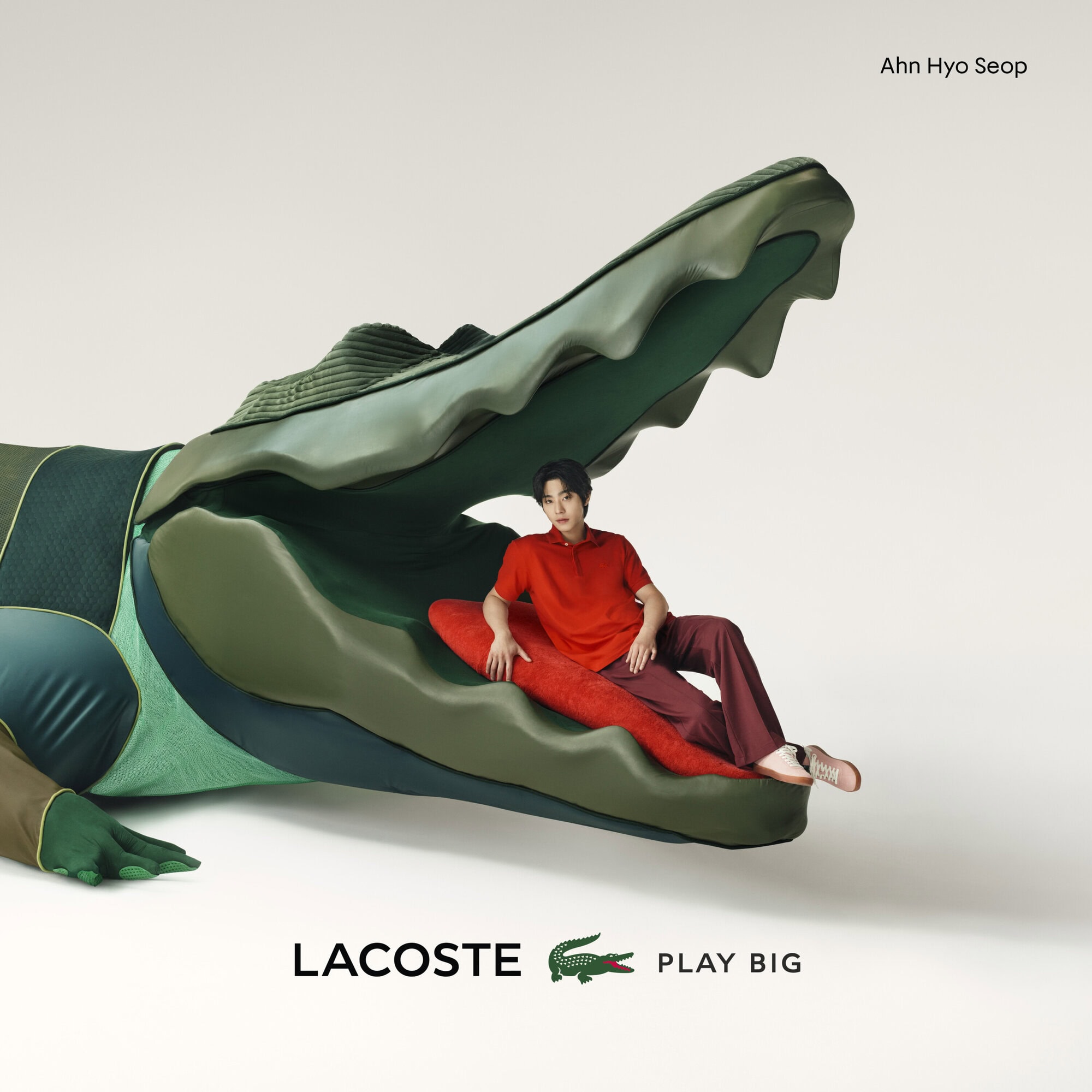 Ahn Hyo Seop posing inside a large green Lacoste alligator head, wearing a red polo shirt and brown trousers against a white backdrop.
