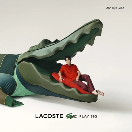 Ahn Hyo Seop posing inside a large green Lacoste alligator head, wearing a red polo shirt and brown trousers against a white backdrop.