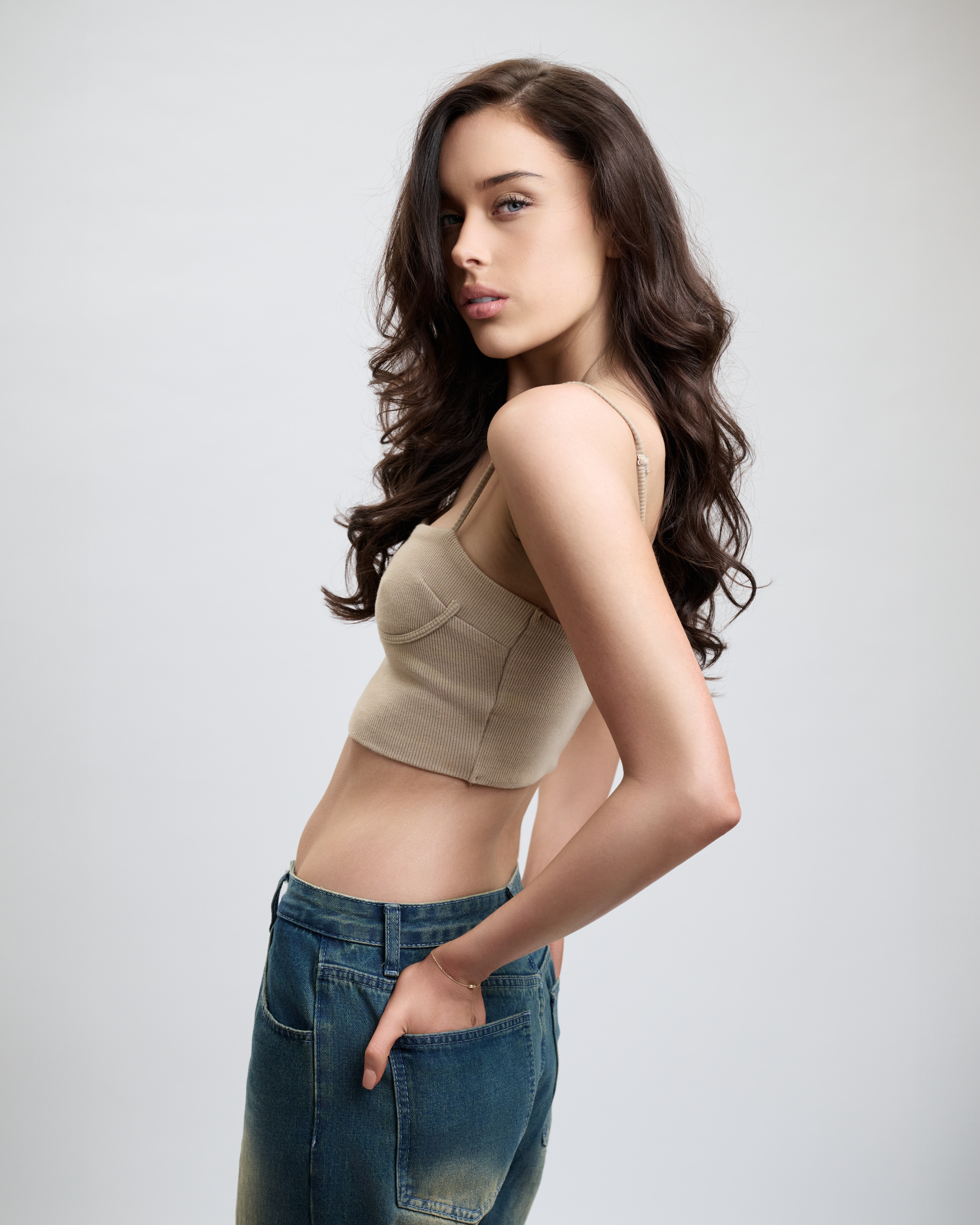 Brunette woman in a crop top and wide-leg jeans poses in a studio, looking over her shoulder with a neutral expression.