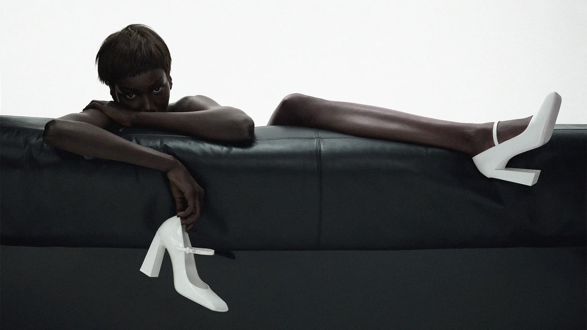 An African-American woman reclines on a black leather couch, gracefully holding a white high-heel shoe, embodying sleek elegance.