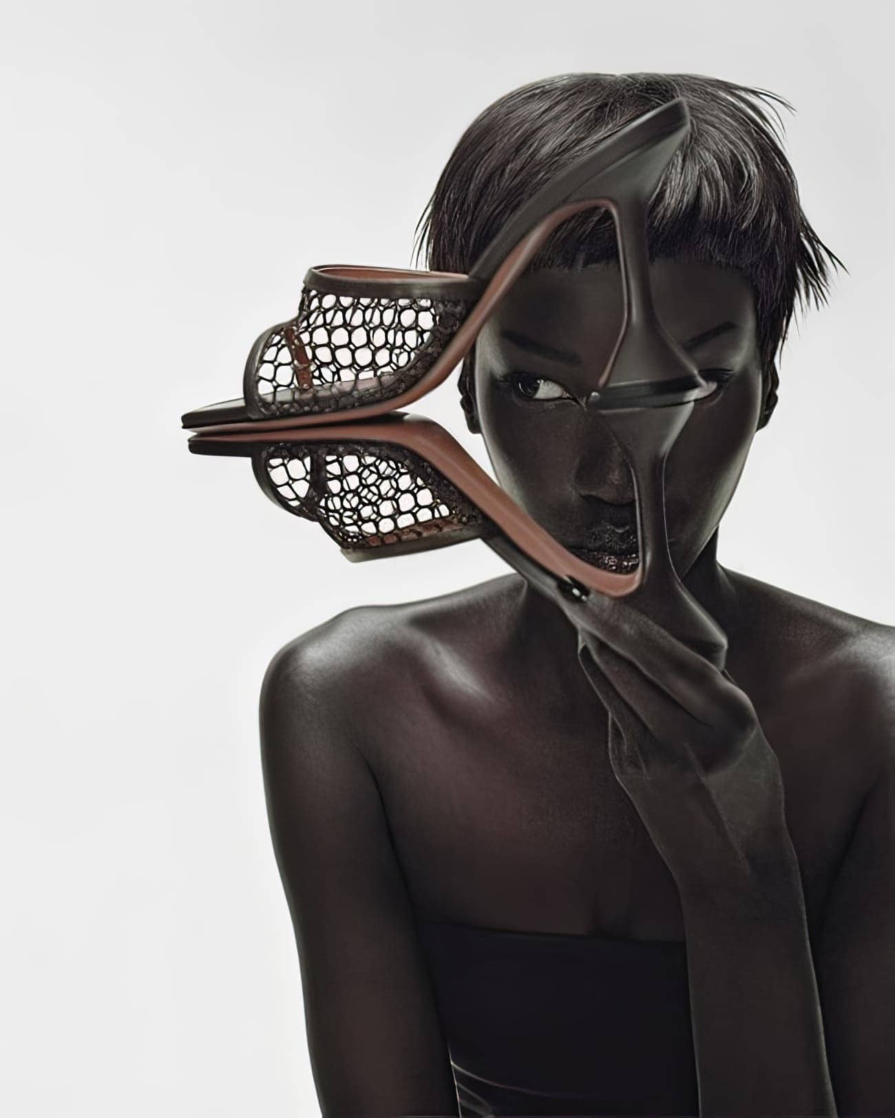 Dark-skinned woman holds high-heeled mule shoe over eye against white backdrop in fashion-forward portrait emphasizing visual unity.