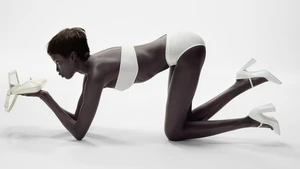 Anok Yai on all fours in white underwear and white heels holding a white handbag against a white background