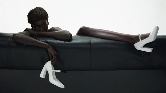 Model Anok Yai reclining on a dark leather couch holding white heels for Amina Muaddi Spring 2024 campaign