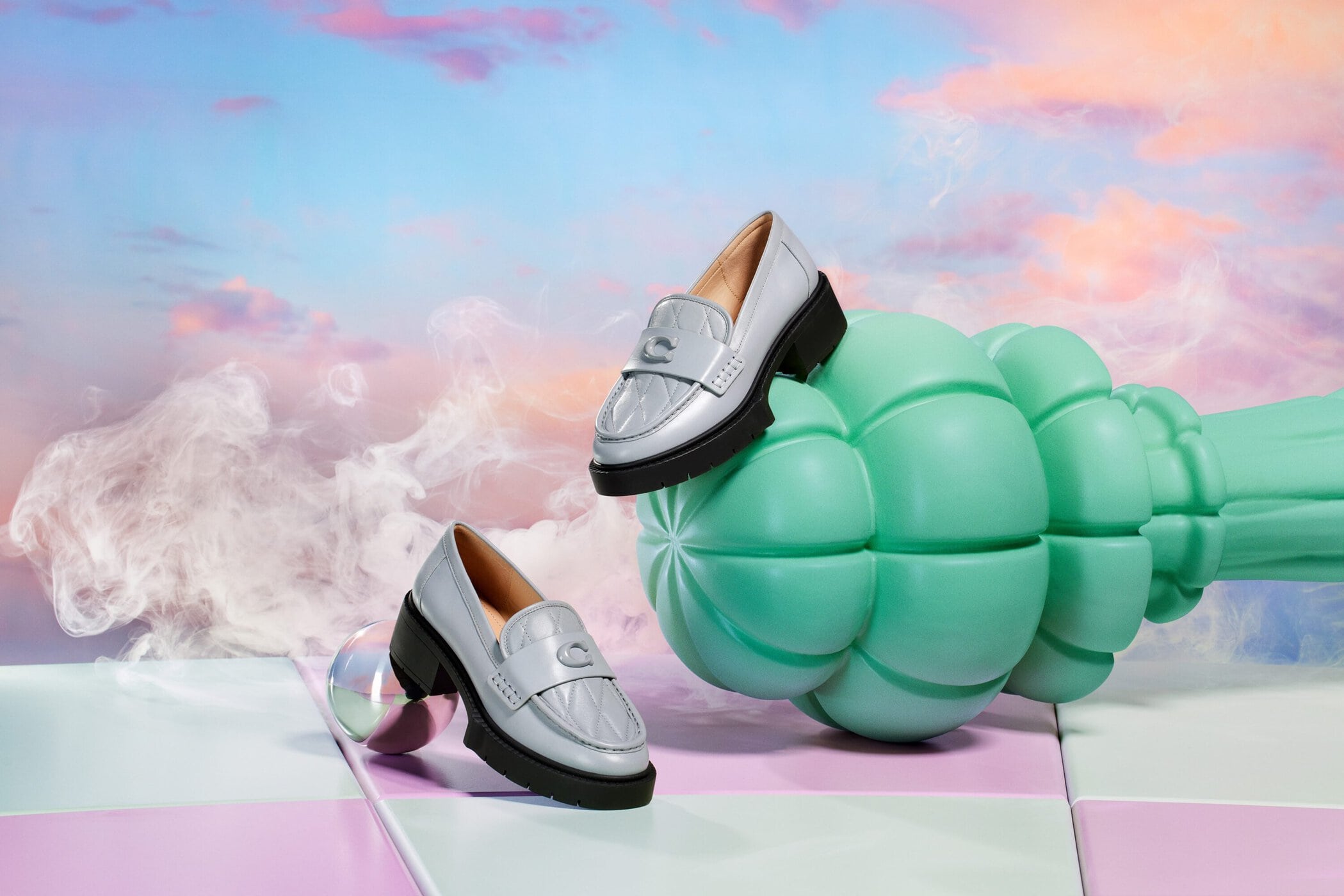 Fashion product shot featuring two silver leather loafers with black soles, displayed on colorful tiles against a cloudy sky background.