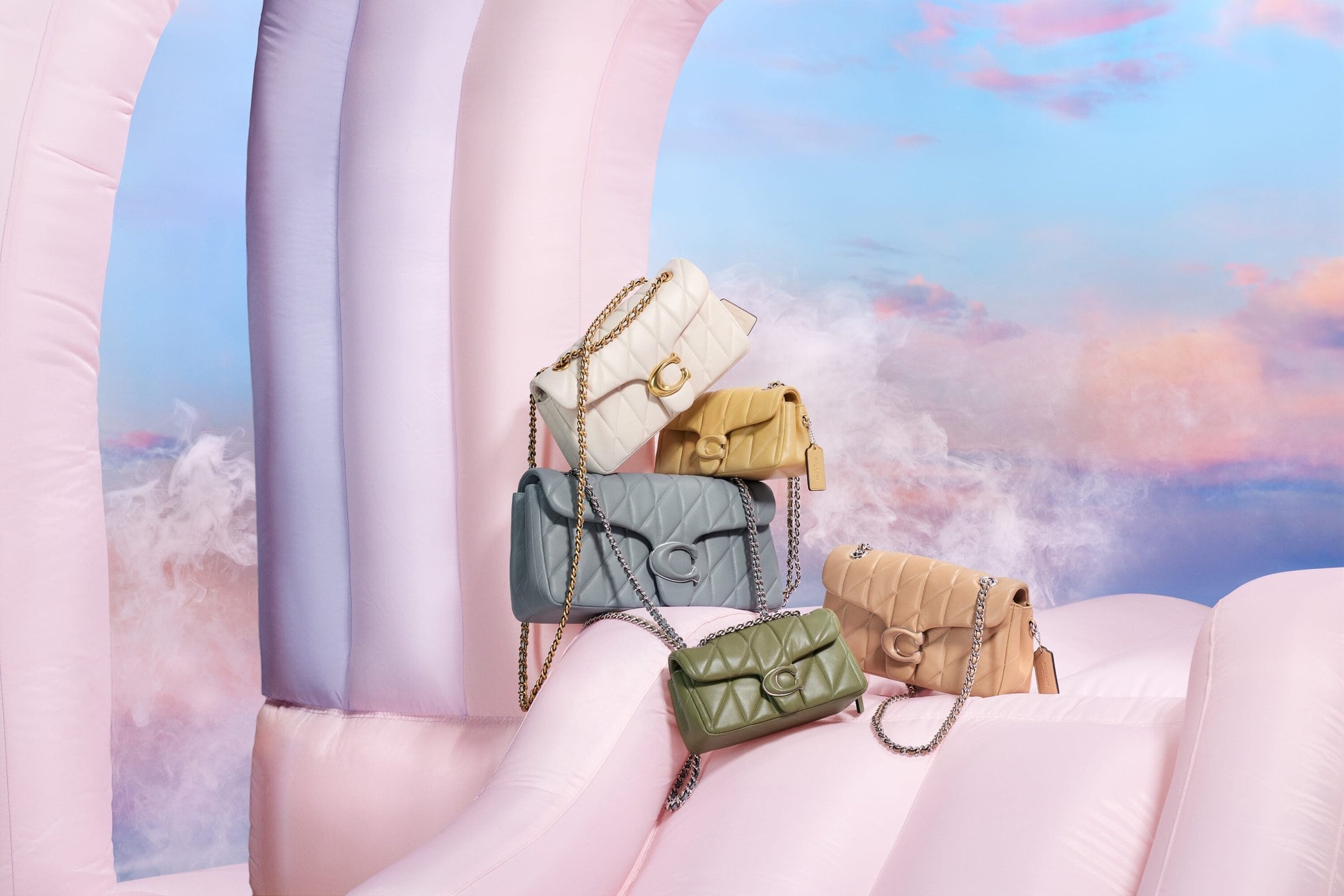 Colorful Coach bags stacked on pink inflatable structure against a cloudy sky, showcasing a playful fashion product display.