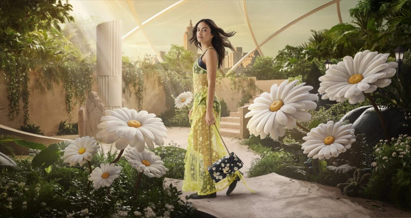 A woman in a floral dress stands on a stone path, surrounded by oversized daisies in an indoor garden with cityscape backdrop.