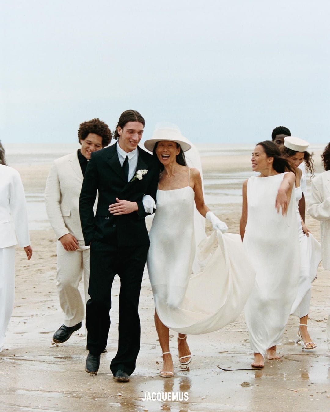 Bride and groom walk on the beach with wedding party; the bride wears a white dress, and the groom wears a black suit in daylight.