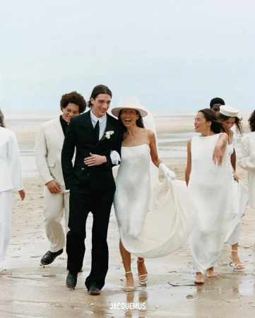 Bride and groom walk on the beach with wedding party; the bride wears a white dress, and the groom wears a black suit in daylight.