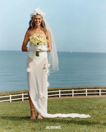 Elegant bride in white gown with floral crown and veil, holding a bouquet. Coastal outdoor wedding on a sunny day, featuring Jacquemus branding.