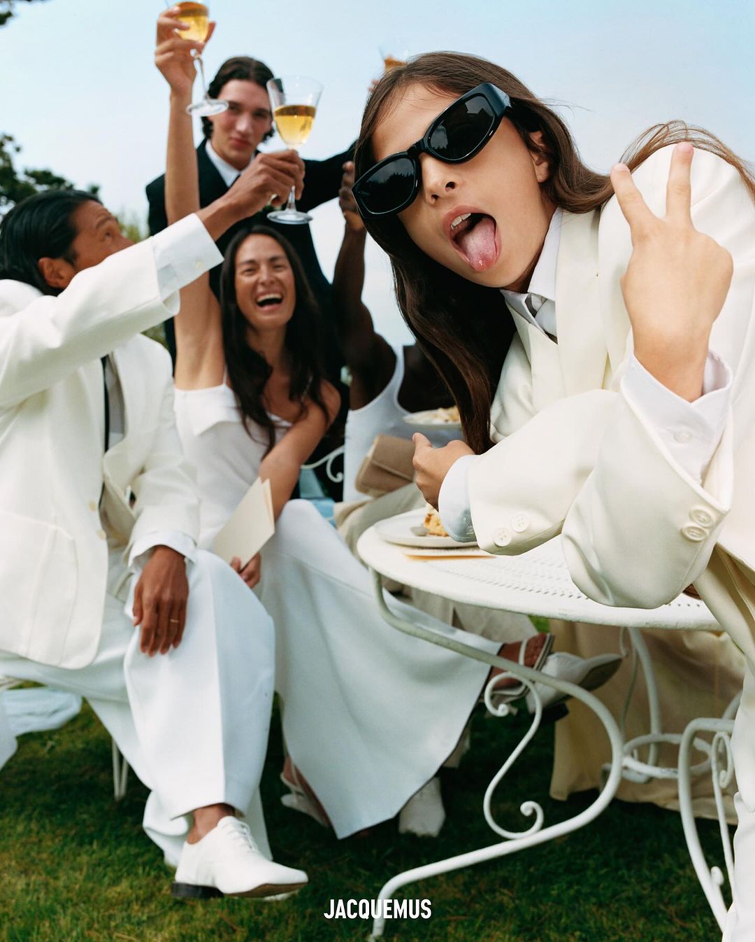 A group of stylish people in white outfits celebrate outdoors with champagne flutes, a woman making a peace sign and sticking her tongue out.