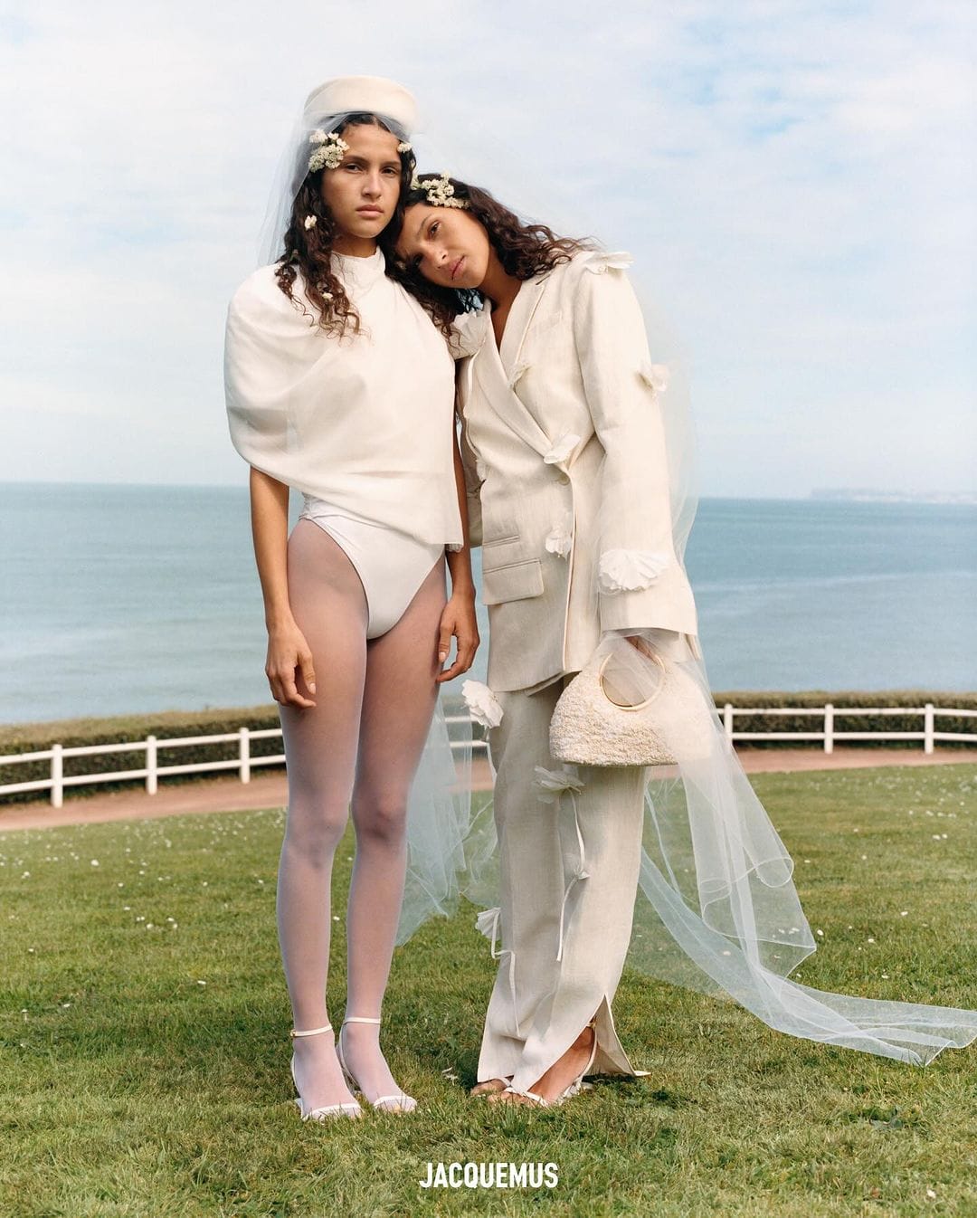 Two models pose on a grassy hill overlooking the ocean in a fashion shot. One wears a bridal-inspired outfit, while the other leans on her.