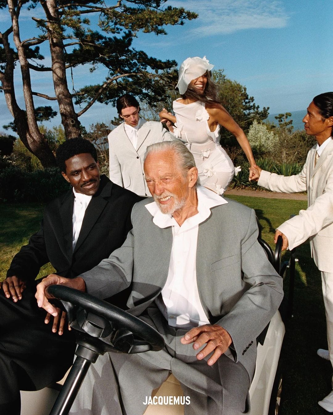 Elderly man driving a golf cart, smiling, with a group of people standing around on a grassy lawn under a clear blue sky.
